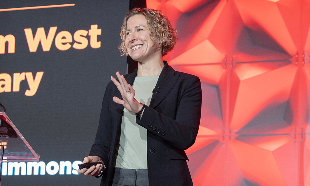 Plenary presenter in a dark suit and with short blonde hair giving a presentation on stage with a red geometric background and the text Quantum West partially visible on the screen behind them.