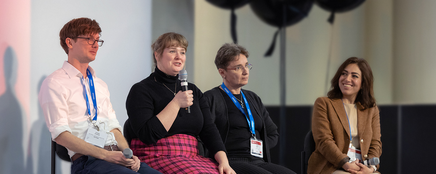 Four people sit in chairs on a stage. The person second from left is holding a mic.