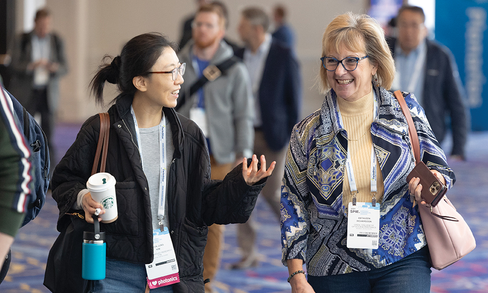Two SPIE conference attendees walking through an event hallway, wearing badges and carrying personal items. One attendee holds a coffee cup and a water bottle while engaged in conversation. Other participants are visible in the background.
