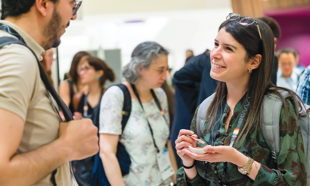 At an SPIE Member Lounge, several attendees stand and converse. In the foreground, one attendee wearing a conference badge and backpack holds a small plate of food while speaking with another attendee carrying a shoulder bag. Additional attendees with lanyards and backpacks fill the background.