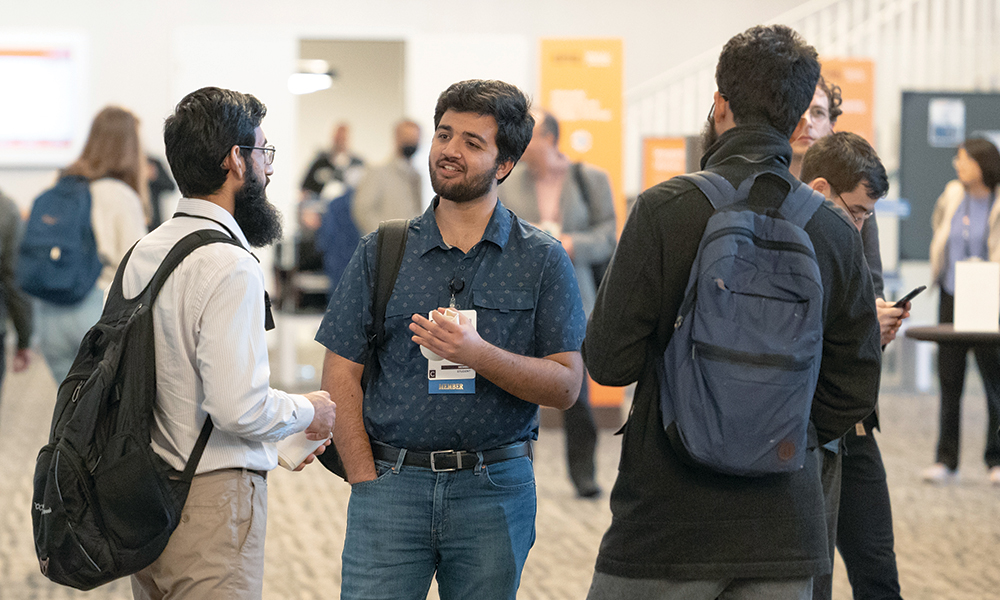 Three SPIE conference attendees standing and talking in a networking area. They are wearing event badges and carrying backpacks. In the background, other participants and informational signage are visible.