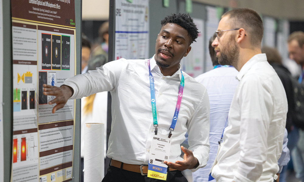 SPIE conference attendee presenting a research poster to another participant in a poster session area. The presenter is pointing to a section of the poster, which includes diagrams and text, while wearing an event badge and lanyard. Other posters and attendees are visible in the background.