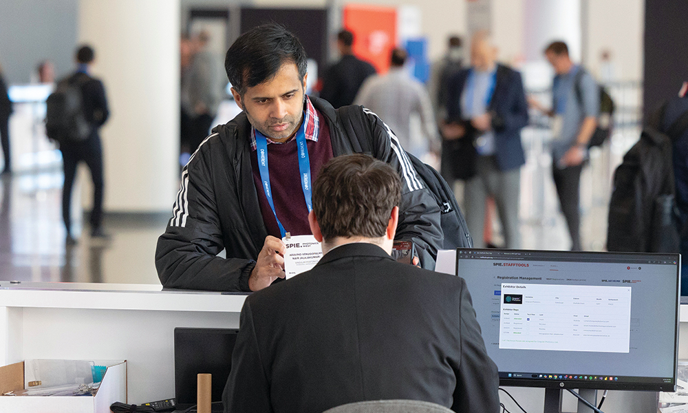 SPIE conference attendee at a registration or information desk interacting with staff. A computer monitor displaying conference information is visible on the desk, along with office supplies. Other attendees are walking in the background.