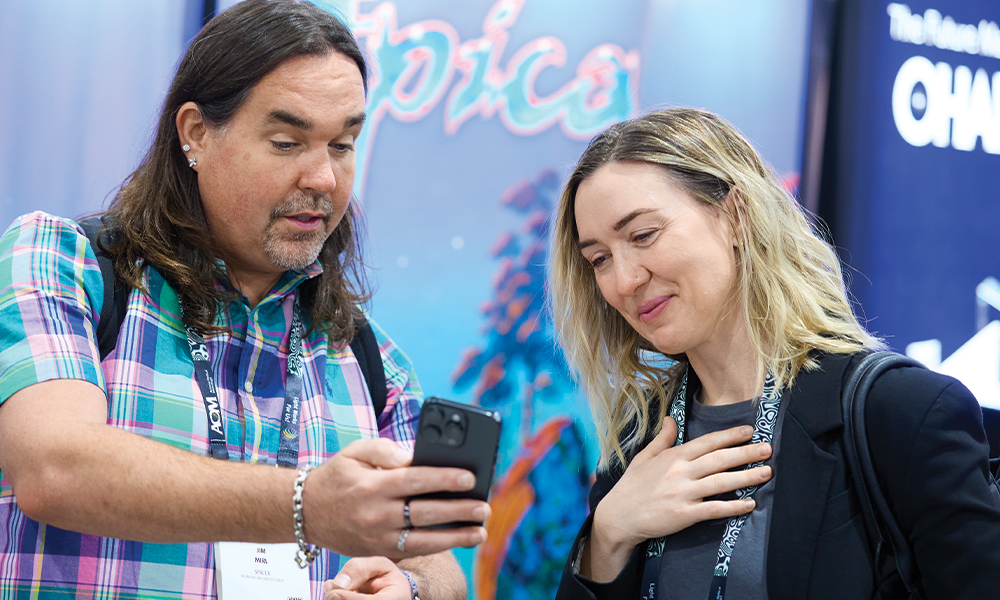 Two SPIE conference attendees standing together, looking at a smartphone screen. Both are wearing event lanyards and badges, and the background includes colorful signage and displays.