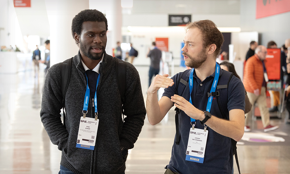 Two individuals walk and converse in a brightly lit convention center during a professional event. Both wear SPIE lanyards with QR-coded badges. One wears a dark sweater over a collared shirt with hands in pockets, while the other gestures with hands and wears a navy blue polo shirt. Other attendees are visible in the background.