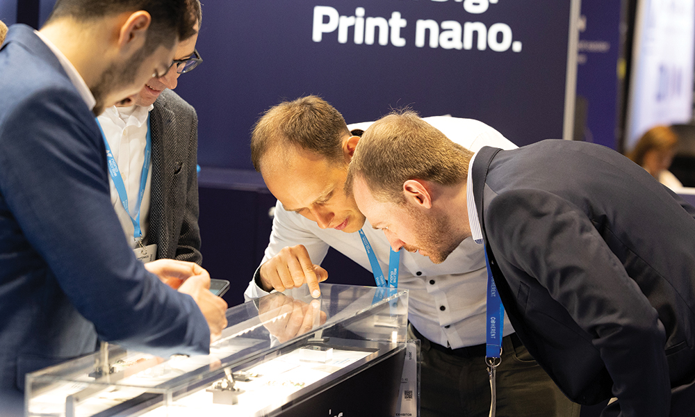 Three individuals closely examining items displayed in a glass case at Photonics West Exhibition, with a dark blue wall in the background featuring partially visible text that reads print nano.