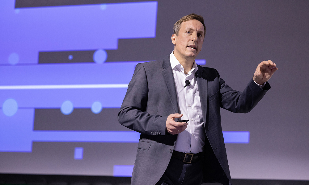 Person in a business suit giving a presentation on stage, holding a small device, with a microphone clipped to the shirt and a large screen displaying abstract geometric shapes in the background.