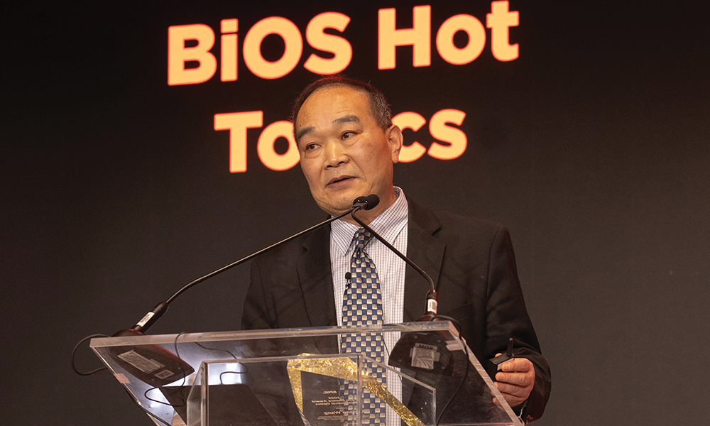 Ruikang Wang, the 2025 recipient of the SPIE Britton Chance Biomedical Optics Award, addresses an audience at Photonics West 2025. He wears a dark suit and tie, stands behind a clear podium with "BiOS Hot Topics" on the screen behind him. 