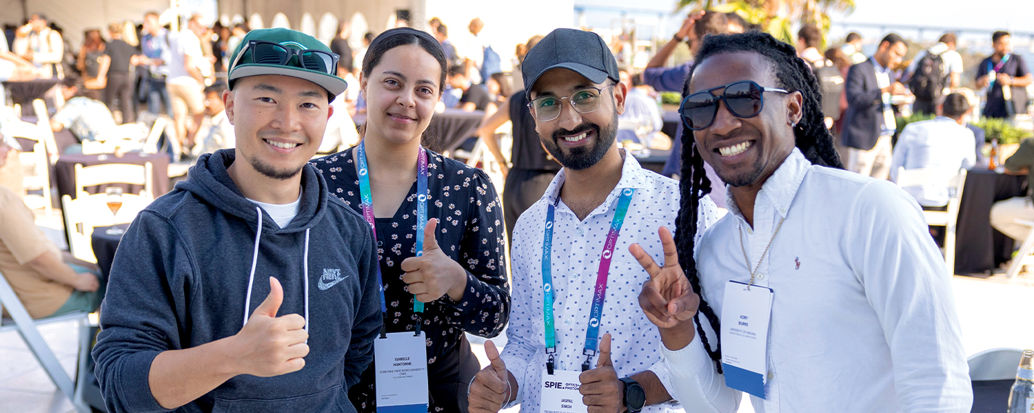 Group of SPIE conference attendees standing outdoors, wearing event badges and casual attire, giving thumbs-up gestures. Other participants and tables are visible in the background, creating a lively networking atmosphere.