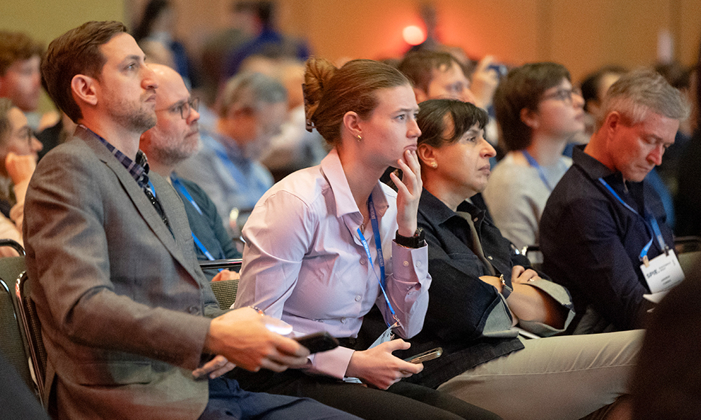 Conference attendees sit closely together in the audience, wearing business‑casual attire and event badges, attentively listening while holding notebooks or mobile devices in a warmly lit indoor venue.