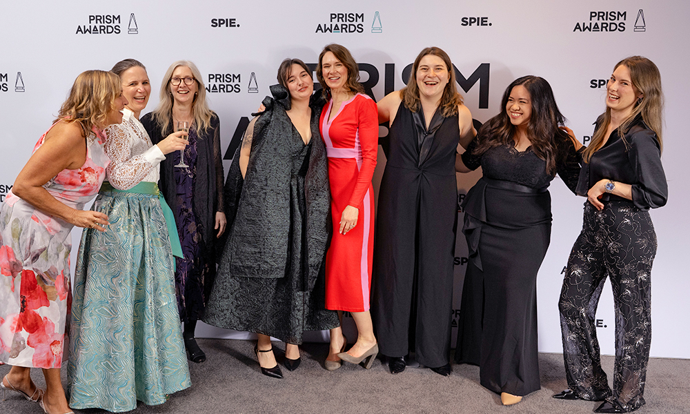 A group of women pose together on a red carpet in front of a PRISM Awards and SPIE step‑and‑repeat backdrop, wearing elegant evening attire in black, red, and patterned fabrics, with arms around one another in a celebratory stance.