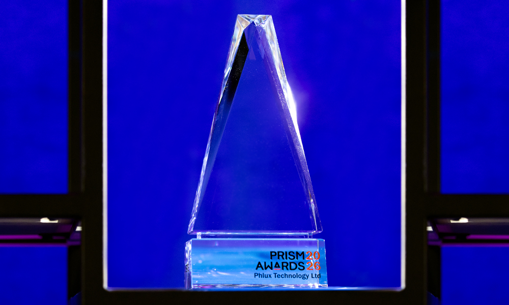 A clear, triangular crystal PRISM Award trophy displayed in a lit showcase against a deep blue background, with an engraved base reading “PRISM Awards 2026"