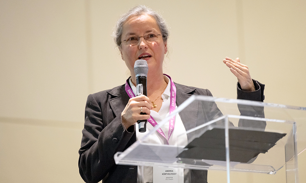 A professional speaker stands at a clear lectern, holding a handheld microphone and gesturing with one hand while presenting at the SPIE AR | VR | MR conference, wearing a blazer, white blouse, and a lanyard.