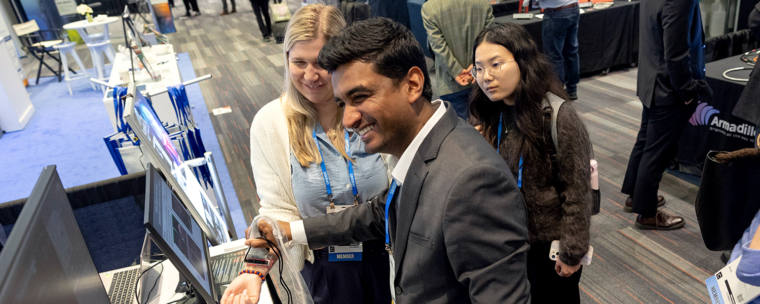At the SPIE BiOS Expo, a technology exhibition, several people gather closely around a biometric demonstration. In the foreground, one person wearing a dark business jacket is holding a small electronic device near a computer monitor, as if demonstrating or scanning something. Two other people stand nearby observing the screen. The group is wearing conference badges on lanyards. The booth includes a flat‑panel display, cables, and a counter surface. In the background, additional booths, signage, and attendees are visible.