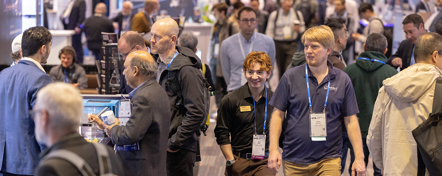 SPIE Photonics West Exhibition shows a crowded technology and industry trades show, as numerous attendees wearing conference badges move through aisles lined with booths and demonstration stations. In the foreground, several people stand near a display counter with electronic equipment and a monitor, while others walk past along the carpeted floor. Booth signage, screens, attendees walking, and product displays are visible throughout the background.