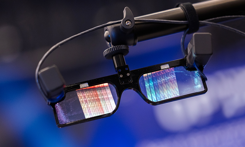 Close-up of smart glasses suspended on a mount, with reflective lenses showing colorful light patterns, set against a softly blurred blue background suggestive of a technology or photonics presentation environment.