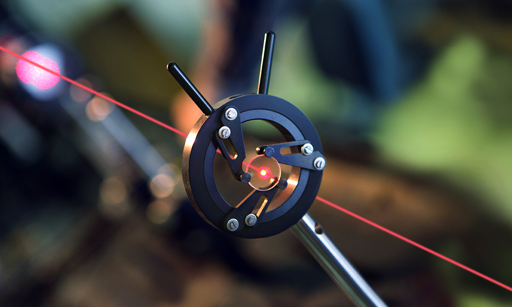 Close-up photograph of a precision optical setup showing a red laser beam passing through the center of a circular black metal alignment mount attached to a tripod or stand. The mount has several visible screws and adjustment arms, with the laser forming a bright red point at the center and a straight beam extending diagonally across the frame.