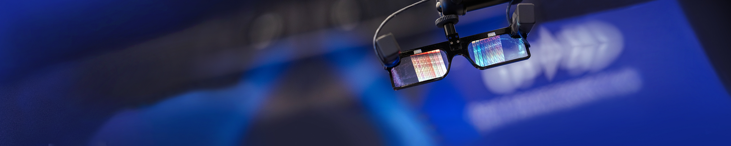 Close-up of smart glasses suspended on a mount, with reflective lenses showing colorful light patterns, set against a softly blurred blue background suggestive of a technology or photonics presentation environment.