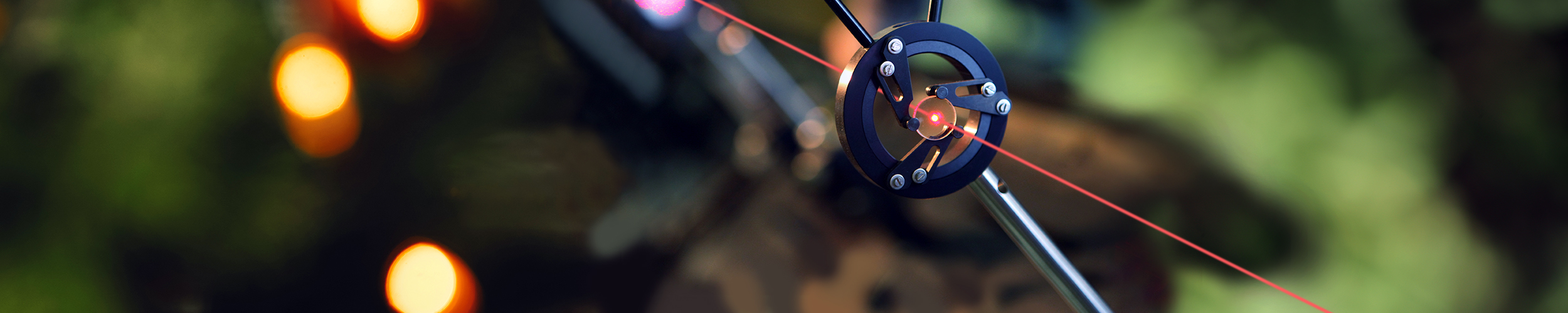 Close-up photograph of a precision optical setup showing a red laser beam passing through the center of a circular black metal alignment mount attached to a tripod or stand. The mount has several visible screws and adjustment arms, with the laser forming a bright red point at the center and a straight beam extending diagonally across the frame.