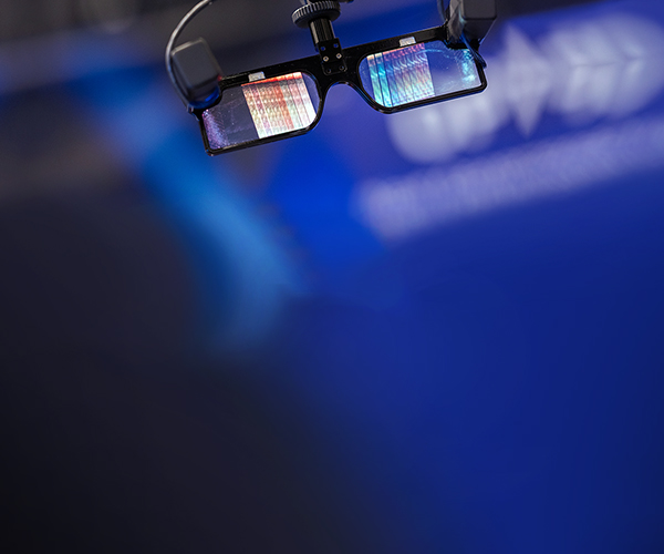 Close-up of smart glasses suspended on a mount, with reflective lenses showing colorful light patterns, set against a softly blurred blue background suggestive of a technology or photonics presentation environment.