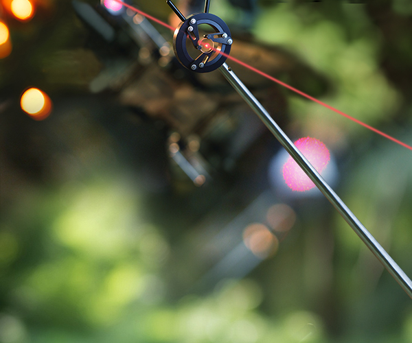 Close-up photograph of a precision optical setup showing a red laser beam passing through the center of a circular black metal alignment mount attached to a tripod or stand. The mount has several visible screws and adjustment arms, with the laser forming a bright red point at the center and a straight beam extending diagonally across the frame.