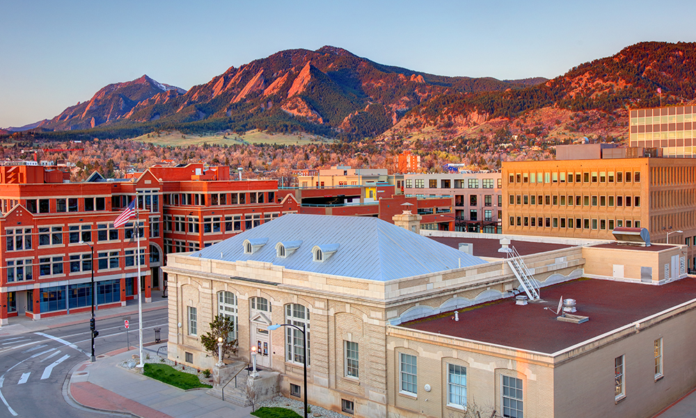 Downtown Boulder, Colorado, showcasing a beige building with a silver roof and red-brick facades, an American flag on a pole, and the Flatirons mountains glowing in warm sunlight under a clear blue sky.