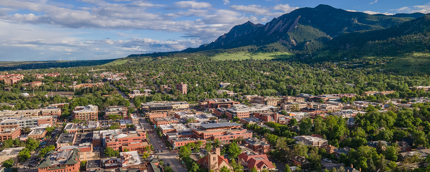 erial view of downtown Boulder, Colorado, with its grid of buildings and tree-lined streets nestled at the base of the jagged Flatirons under a partly cloudy sky.