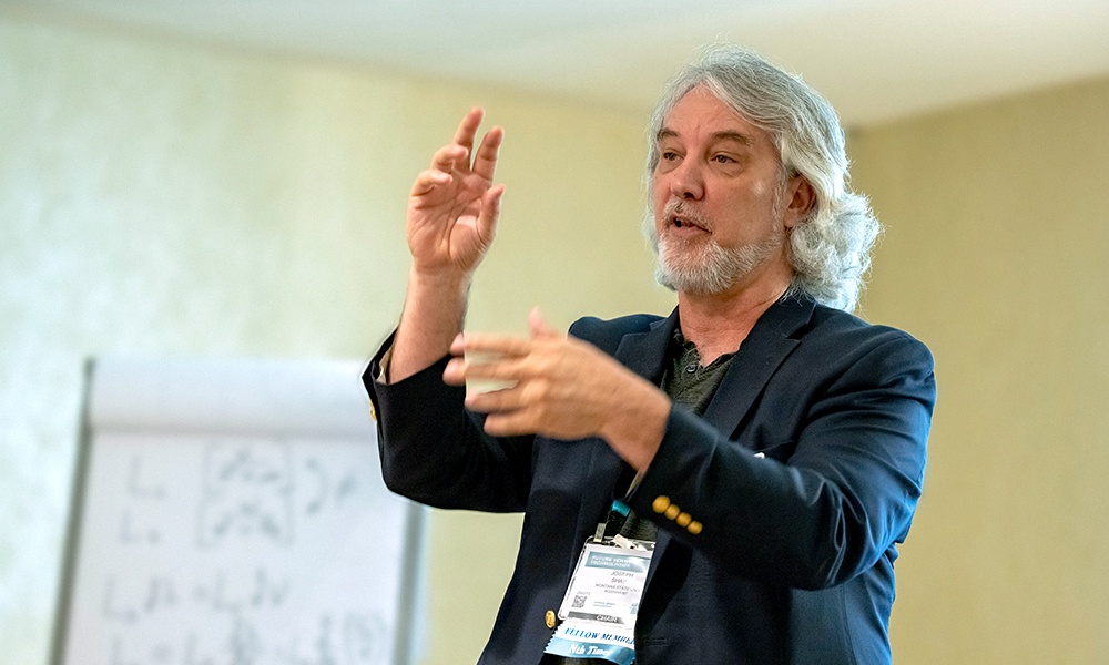 A gray-haired, bearded individual in a dark blazer with gold buttons speaks and gestures with both hands during a presentation. A microphone is clipped to the blazer, and a name badge is visible. Behind them, an easel pad displays partially visible handwritten notes or equations in a conference room setting.