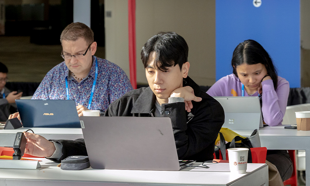 A workspace with several attendees seated at long tables using laptops. In the foreground, one attendee works on a laptop with a coffee cup and notebook nearby. Behind them, two additional attendees sit at the same table with open laptops, wearing conference lanyards and badges. The setting appears to be a designated work or lounge area within a conference venue, with neutral walls, soft lighting, and personal items such as backpacks and cups visible on the tables.