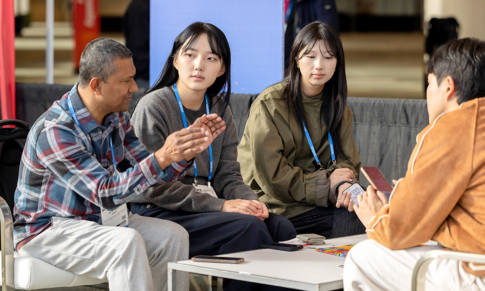 SPIE Member Lounge setting with four attendees seated around a low table, all wearing conference lanyards and badges. One attendee on the left gestures with both hands while speaking, while two attendees in the center sit side by side listening. Another attendee on the right holds a smartphone and faces the group. The background shows a large screen and other conference seating under bright indoor lighting, suggesting an informal professional networking conversation.