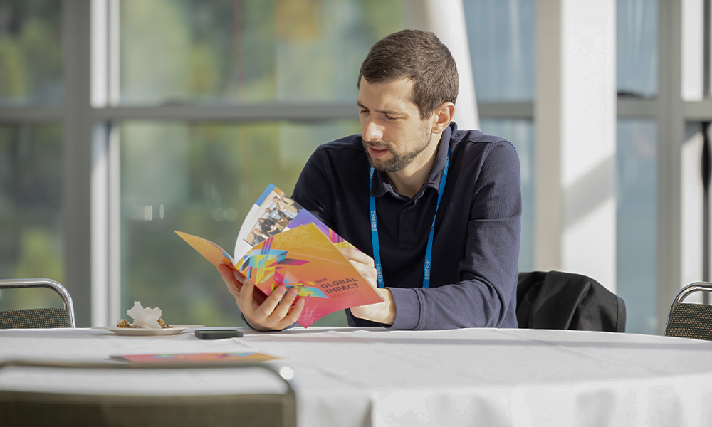 Natural light streams through large windows. An SPIE Member Lounge attendee wearing a conference lanyard sits at a round table and reads a colorful brochure titled “Global Impact.” 