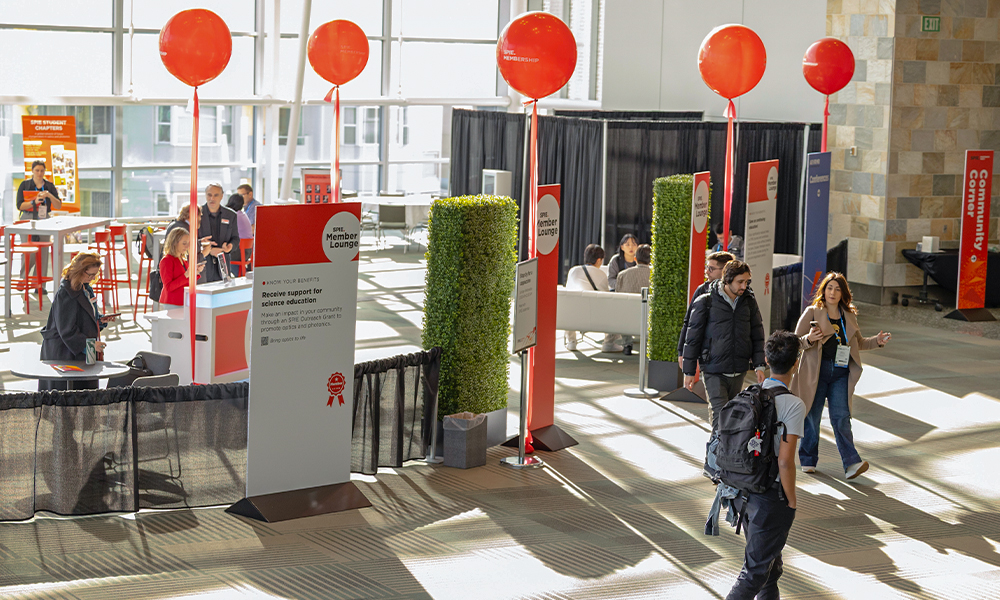 Wide view of the SPIE Member Lounge inside a bright convention center, featuring red SPIE‑branded balloons, vertical lounge signage, and check‑in counters. Attendees with conference badges walk through and gather in seating areas, with partitions, plants, and lounge furniture arranged in an open, welcoming layout illuminated by large windows and natural light.