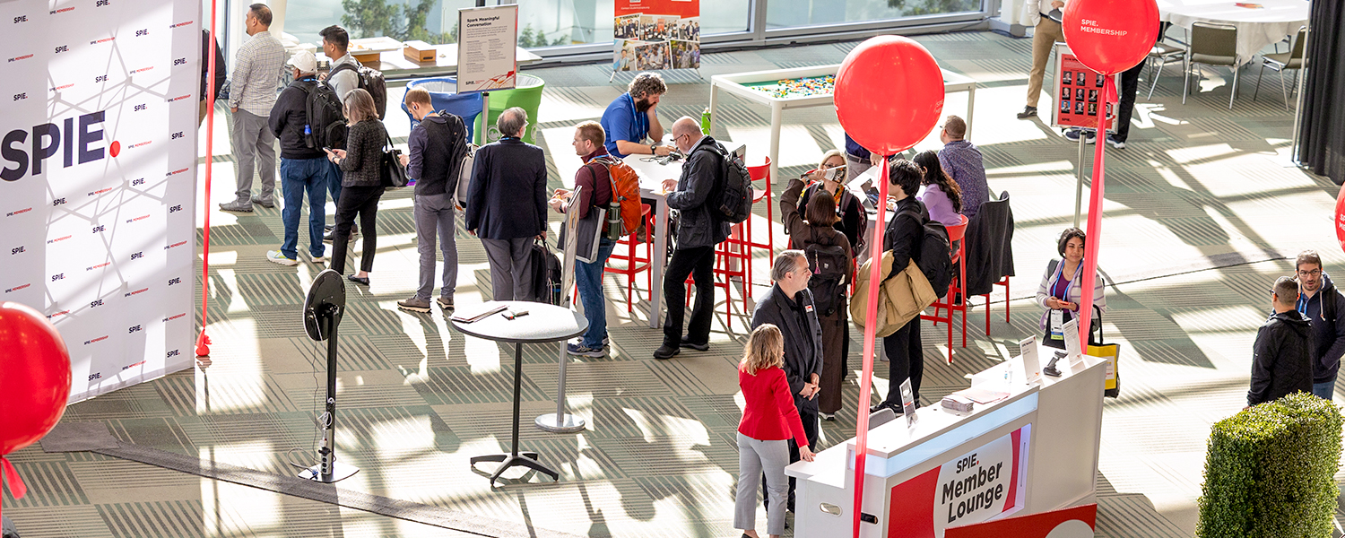 Wide overhead view of a busy SPIE Member Lounge inside a bright convention center. Attendees wearing conference badges gather around high‑top tables and the Member Lounge check‑in counter. Red SPIE‑branded balloons rise above the space, while a step‑and‑repeat SPIE backdrop stands on the left. Sunlight streams through large windows, illuminating seating areas, informational displays, and clusters of attendees engaged in conversation.