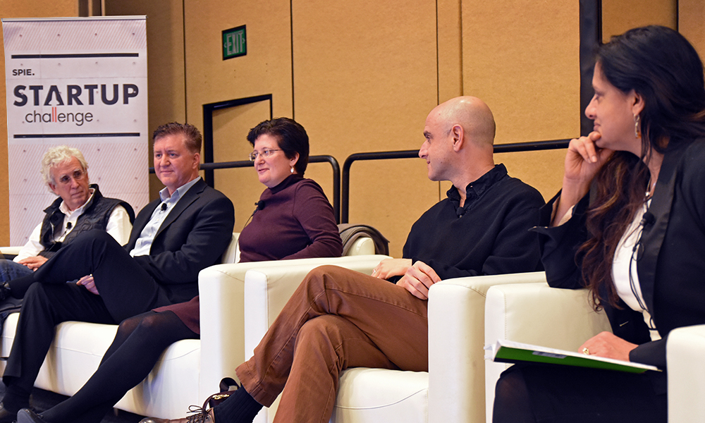 Panel of five individuals seated on white chairs during the SPIE Startup Challenge event, with a banner and beige wall in the background.