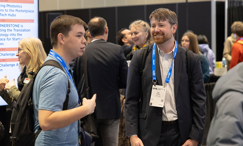 Group of people at a conference or networking event, with two individuals in the foreground conversing while wearing name badges and lanyards; background features other attendees and a large screen displaying text about photonics innovations and tech hubs.