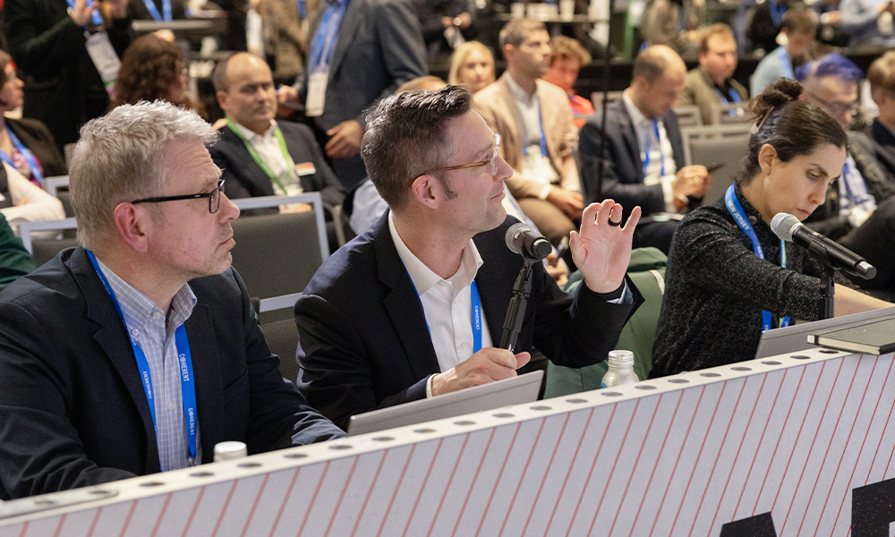 Group of people seated at a conference or meeting, with three individuals in the foreground holding microphones and participating in discussion; background attendees are also seated and facing forward. Faces are blurred for privacy.