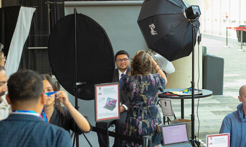An attendee sits for a professional headshot at a free photo station during an SPIE event, with a photographer adjusting the camera and others visible in the background.