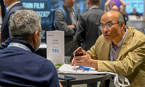 Two people seated at a round table engage in conversation during the SPIE Job Fair, with a 'Free WiFi' sign visible and other participants networking in the background.