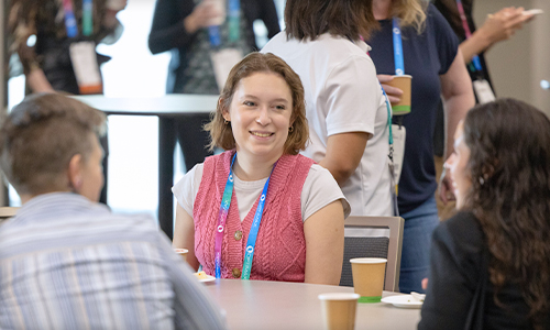 Attendees gather and converse around a table during an SPIE Women in Optics meetup, with coffee cups, conference badges, and a bright, welcoming event setting.