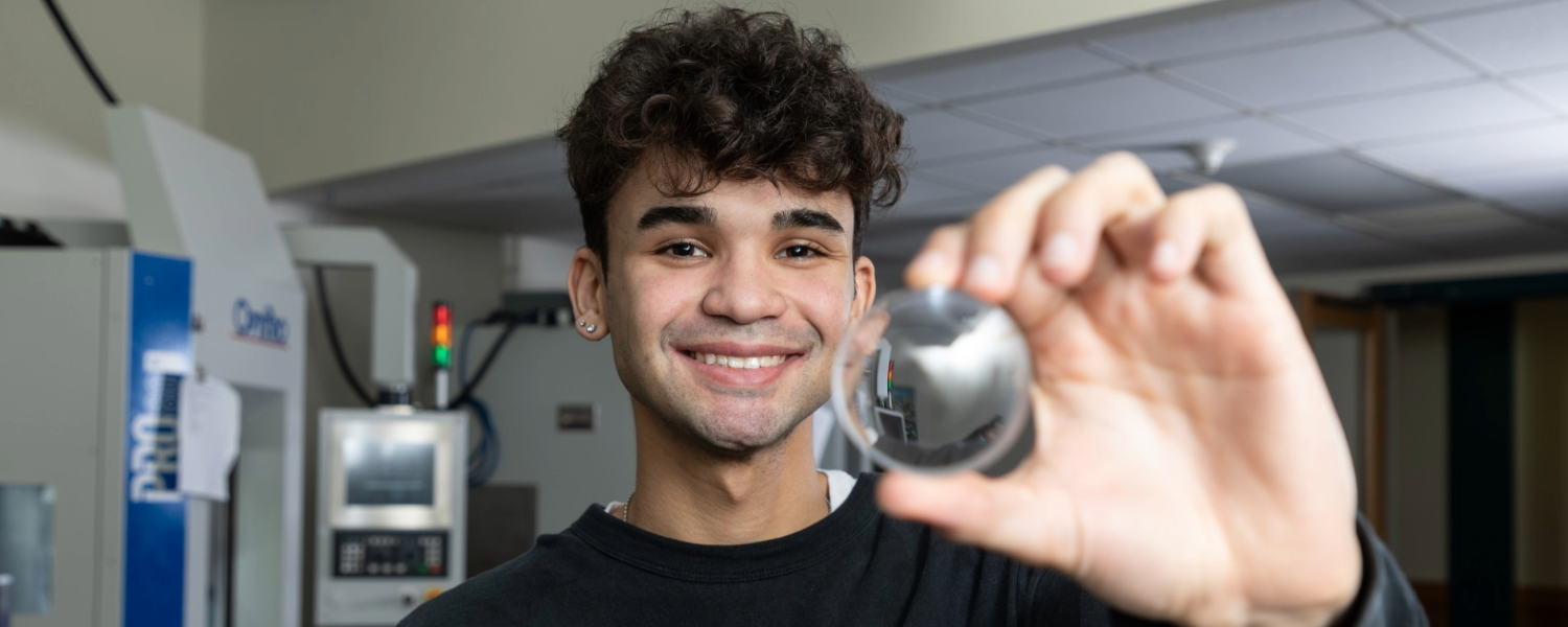 A person holding a shiny lens close to the camera. The background shows a room with machinery and equipment.