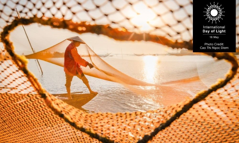The winning photo from the 2025 photo contest, which features a person wearing a traditional conical hat and red clothing walks along a calm body of water at sunrise or sunset, skillfully handling a large fishing net. The golden light casts a warm glow over the scene, highlighting the net's intricate layers and other suspended nets in the distance. Photo by Cao Thi Ngoc Diem.