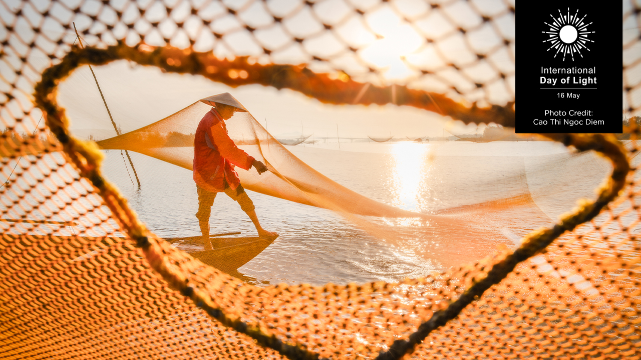 The winning photo from the 2025 photo contest, which features a person wearing a traditional conical hat and red clothing walks along a calm body of water at sunrise or sunset, skillfully handling a large fishing net. The golden light casts a warm glow over the scene, highlighting the net's intricate layers and other suspended nets in the distance. Photo by Cao Thi Ngoc Diem.