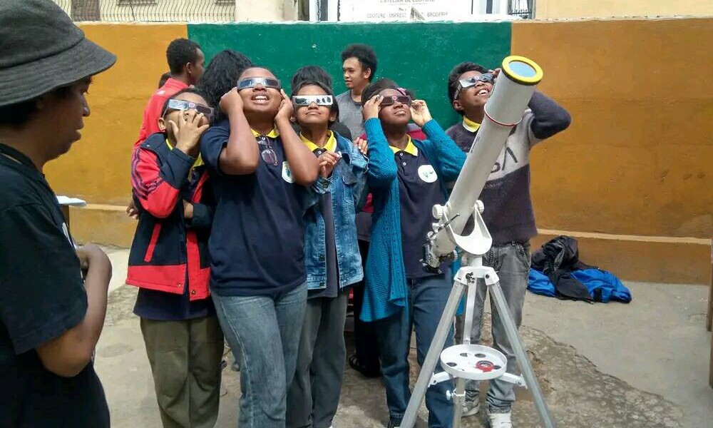 A group of students participating in an outdoor optics outreach activity, taking turns looking through a telescope on a tripod.