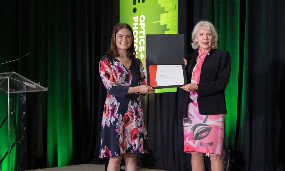 Katherine Koch receives the Women in Optics Scholarship from Donor Janet Fender on a stage at an Optics + Photonics event. They both hold a framed Women in Optics Scholarship certificate against a green-lit backdrop.