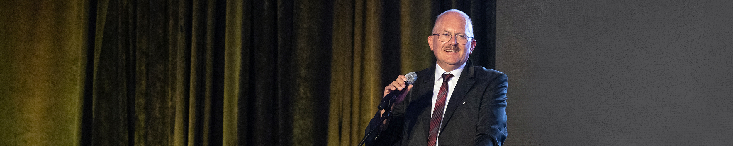 Jürgen Czarske speaks into a microphone after being presented with the 2024 SPIE Dennis Gabor Award in Diffractive Optics at the 2024 Society Awards banquet in San Diego, California. He is wearing a dark suit with a tie and is illuminated by a focused spotlight.