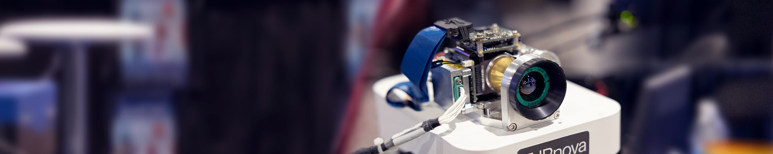A close-up image of a small electronic device with various components, including a lens, circuit board, and cables, mounted on a white rectangular base.