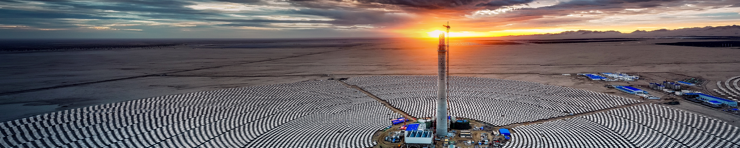 Aerial view of a large solar power plant in a desert landscape at sunset. The plant features a central tower surrounded by thousands of solar panels arranged in concentric circles. The sky is partly cloudy with the sun setting on the horizon, casting an orange glow. Photo credit: Xiaoqing Zou.