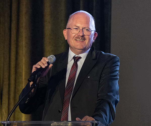 Jürgen Czarske speaks into a microphone after being presented with the 2024 SPIE Dennis Gabor Award in Diffractive Optics at the 2024 Society Awards banquet in San Diego, California. He is wearing a dark suit with a tie and is illuminated by a focused spotlight.