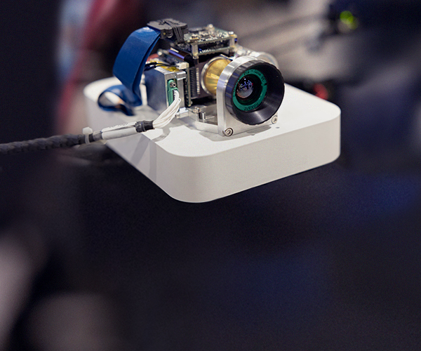 A close-up image of a small electronic device with various components, including a lens, circuit board, and cables, mounted on a white rectangular base.