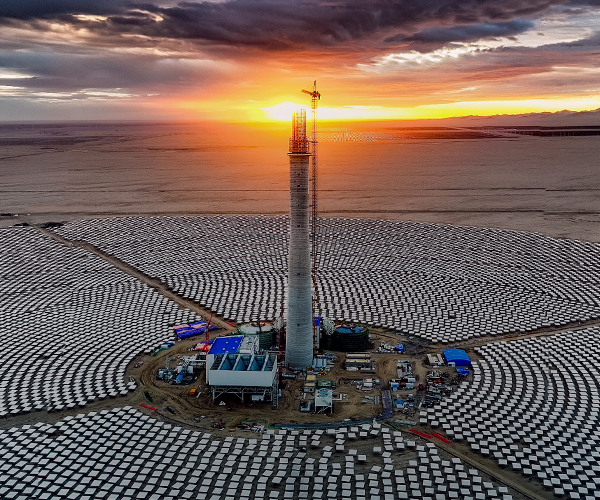 Aerial view of a large solar power plant in a desert landscape at sunset. The plant features a central tower surrounded by thousands of solar panels arranged in concentric circles. The sky is partly cloudy with the sun setting on the horizon, casting an orange glow. Photo credit: Xiaoqing Zou.